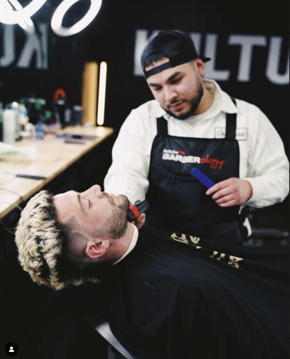 Barber grooming a client's beard with electric clippers in a barbershop.