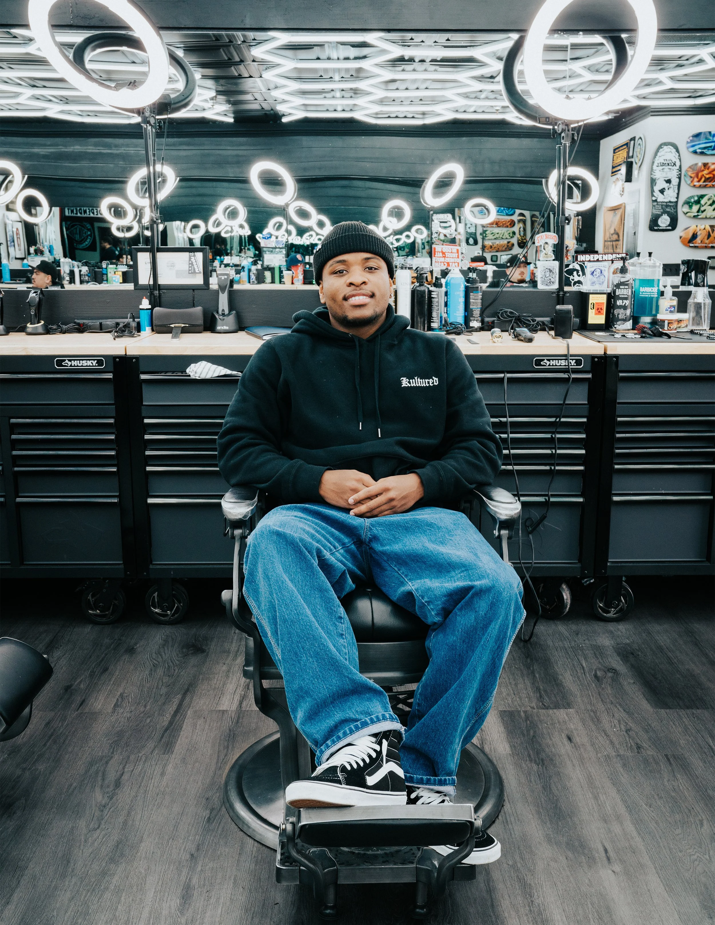A man seated in a barber shop chair behind a station with hair styling products, mirrors, and barber tools, with a modern, well-lit interior featuring circular light fixtures on the ceiling.