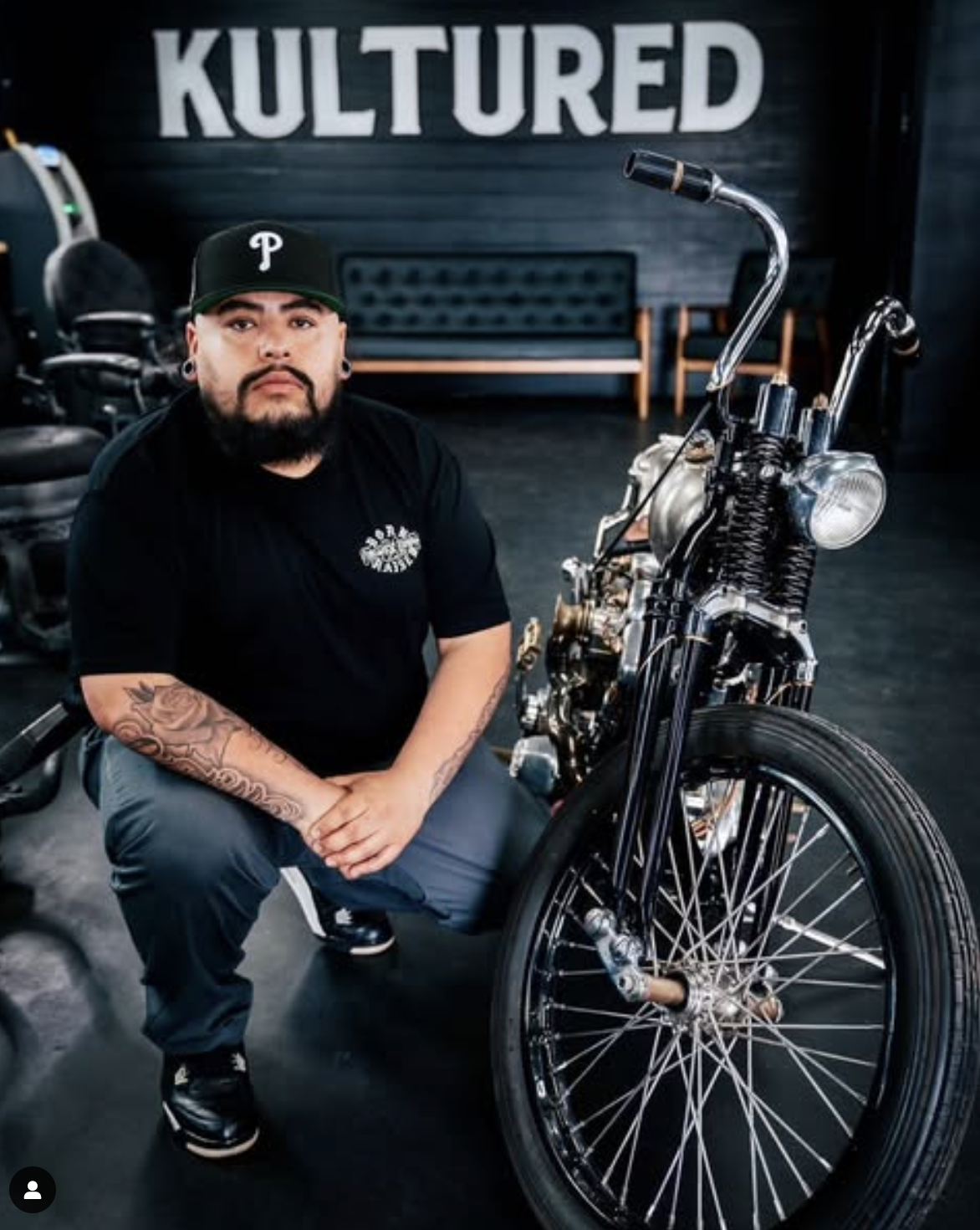 Person squatting next to customized motorcycle in a room with 'Kultured' sign above.