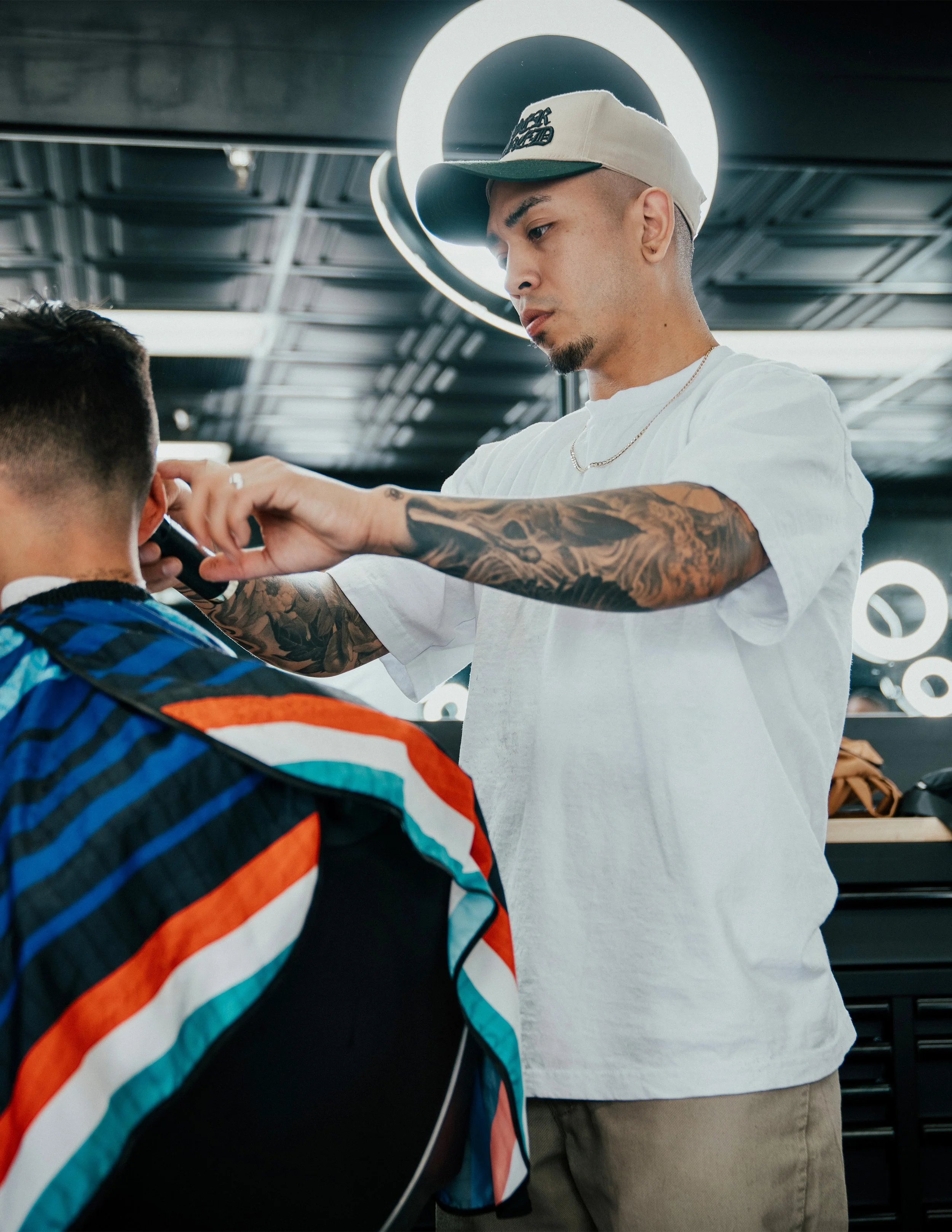 Person sitting outdoors wearing a green cap, white t-shirt, and blue jeans, with tattooed arms.