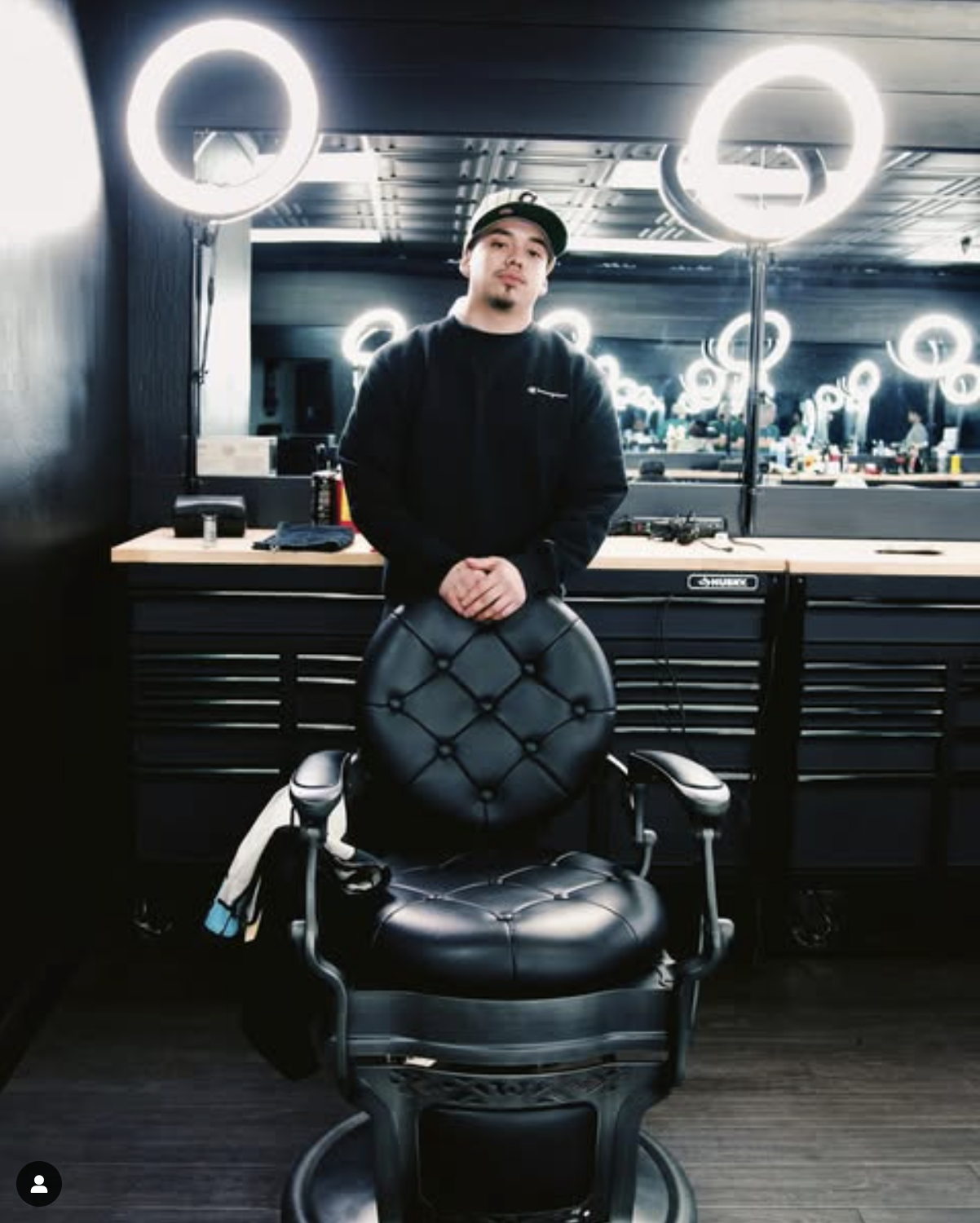 Barber standing behind a black leather salon chair in a modern barbershop with ring lights and mirrors.