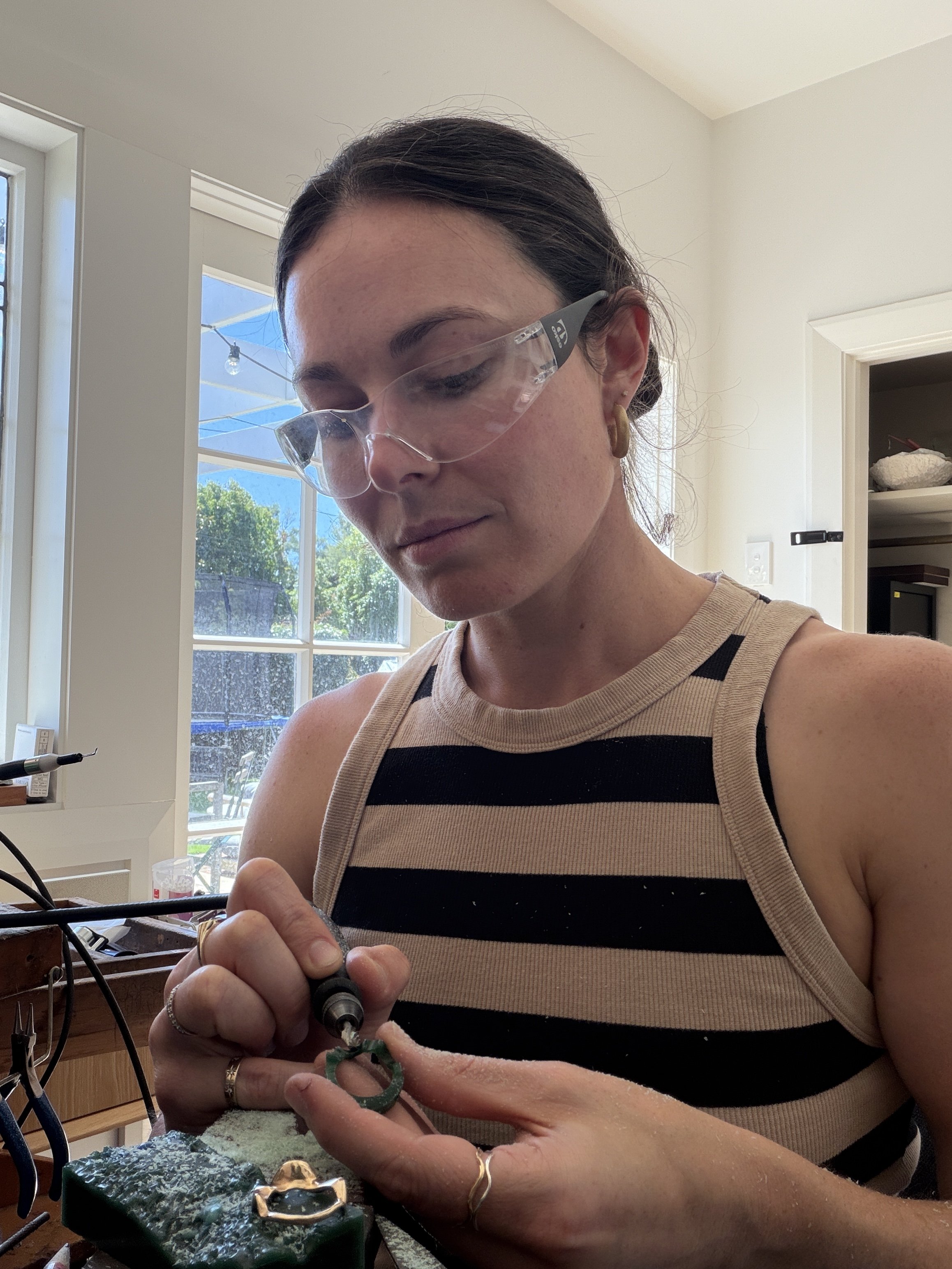 Woman with dark hair wearing safety glasses and a striped tank top working on jewelry with tools at a workbench near a large window.