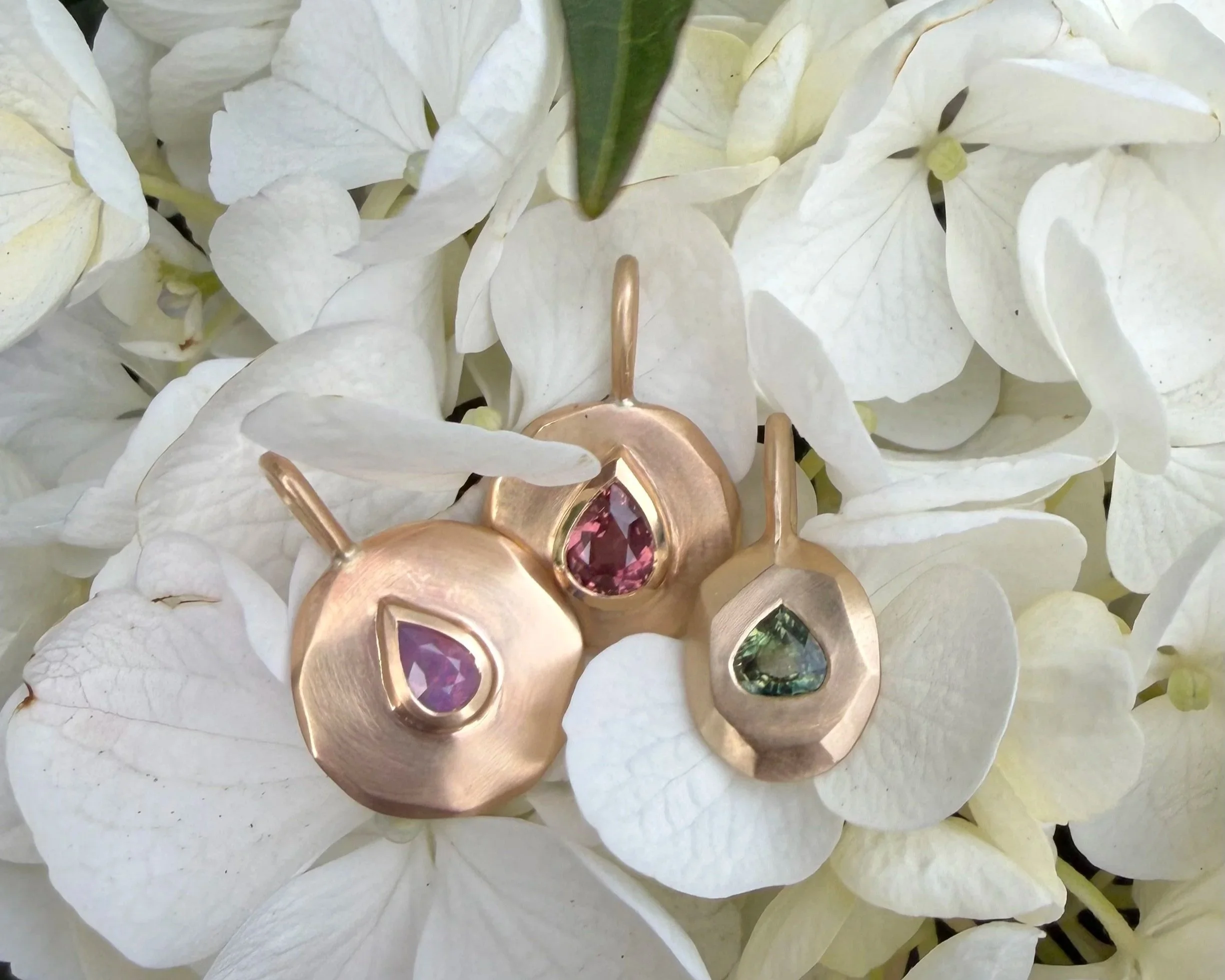 Three gold pendants with colored gemstones resting on white hydrangea flowers.
