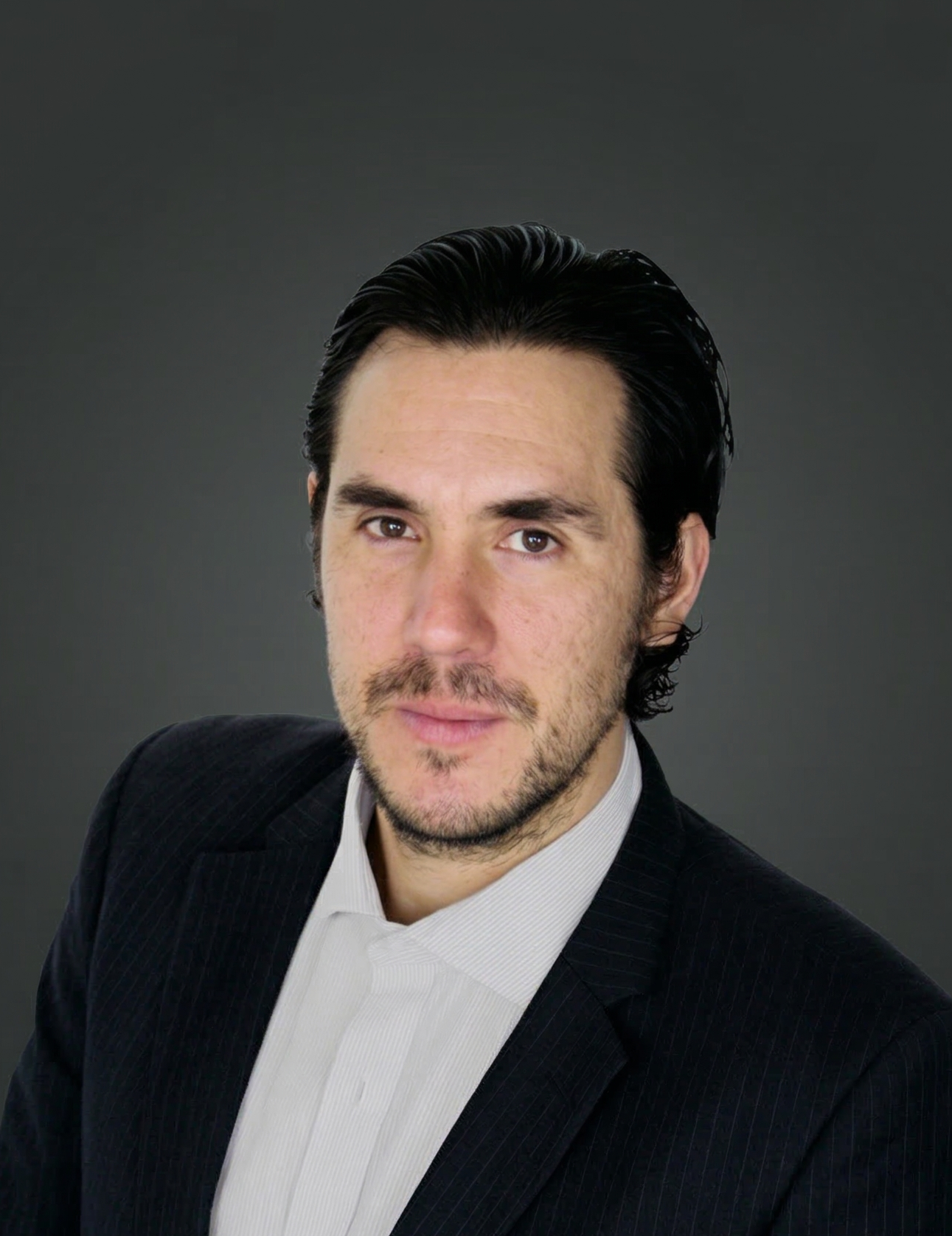 A man with dark, slicked-back hair, wearing a black pinstripe suit and white shirt, against a dark grey background.