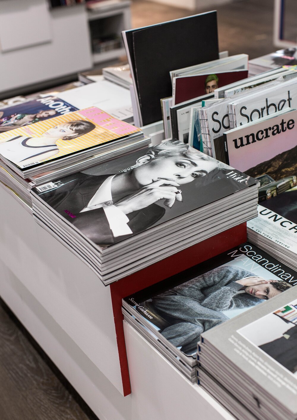 A display of various magazines in a bookstore or magazine shop, including fashion, art, and culture magazines, with a prominent black and white Vogue magazine featuring a woman with short blonde hair.