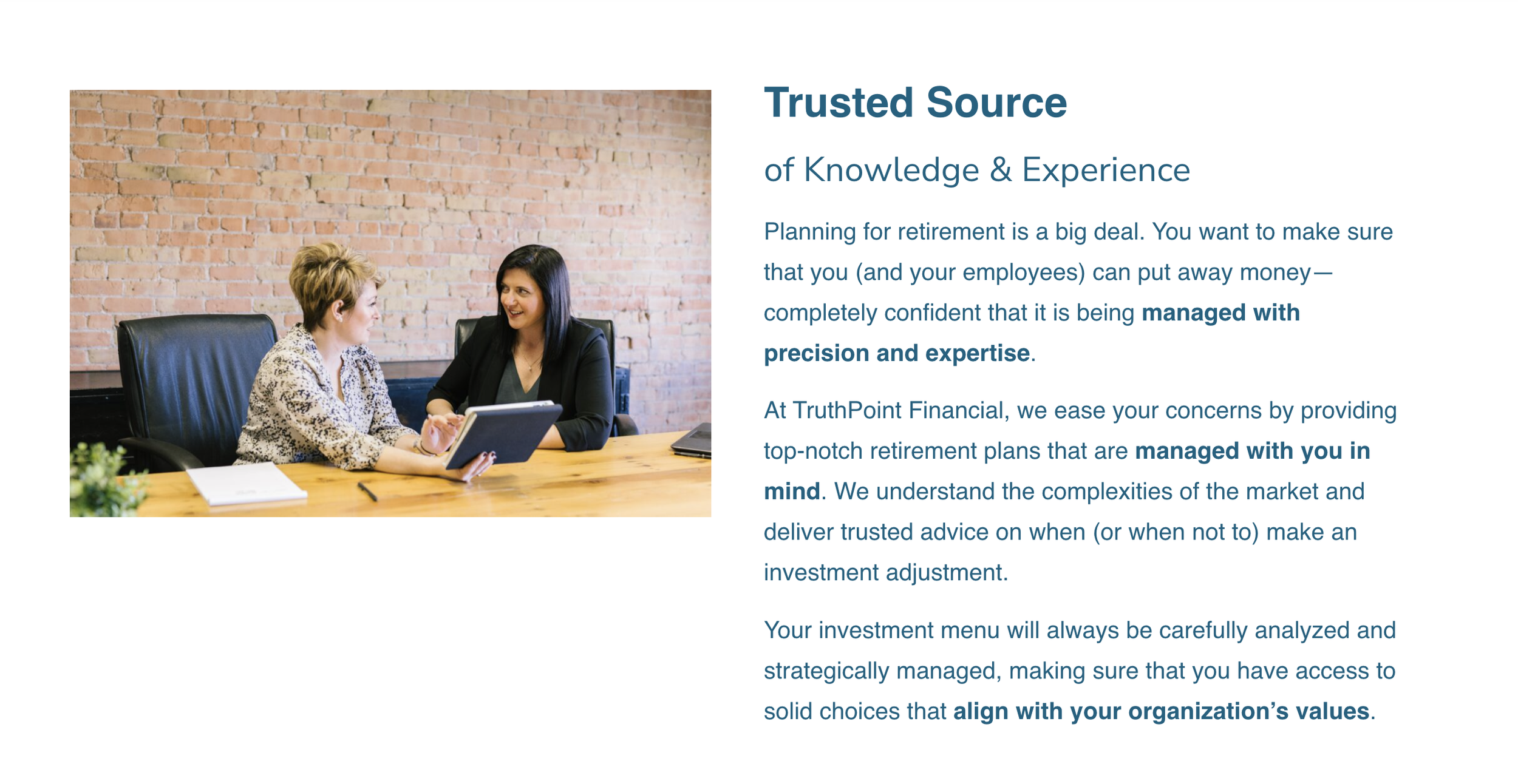 Two women having a discussion in a modern office with an exposed brick wall, one holding a tablet and the other looking at it, sitting at a wooden table.