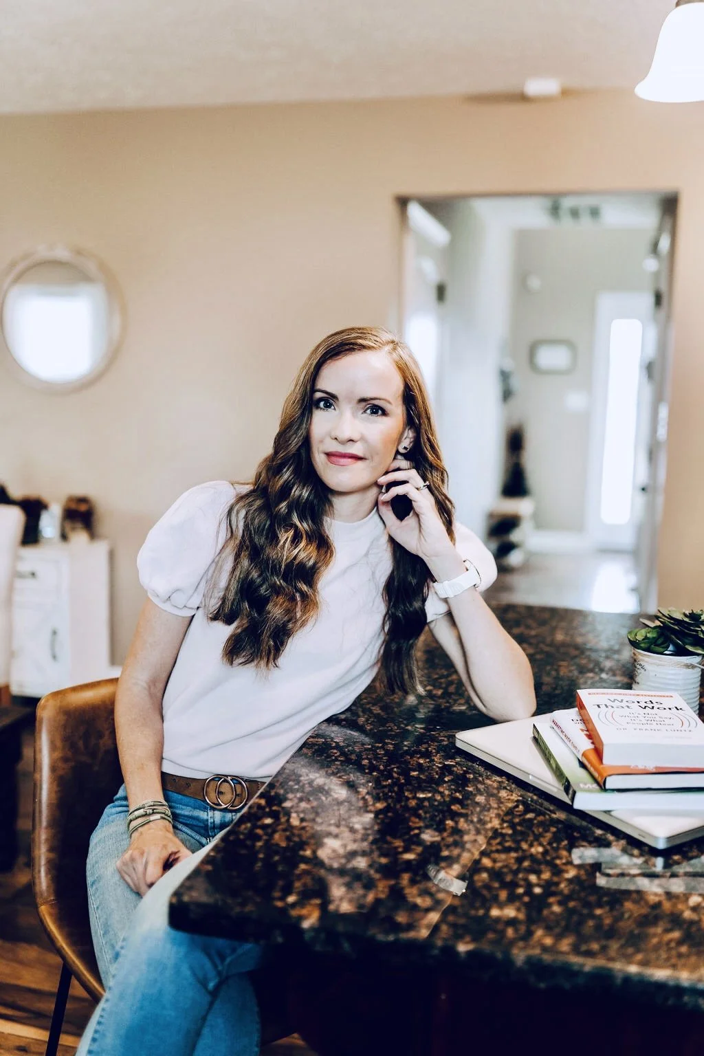 A woman with long wavy brown hair, wearing a white t-shirt, sits at a kitchen counter with her elbow resting on the counter, looking at the camera. There are books and a plant on the counter, and a doorway in the background.
