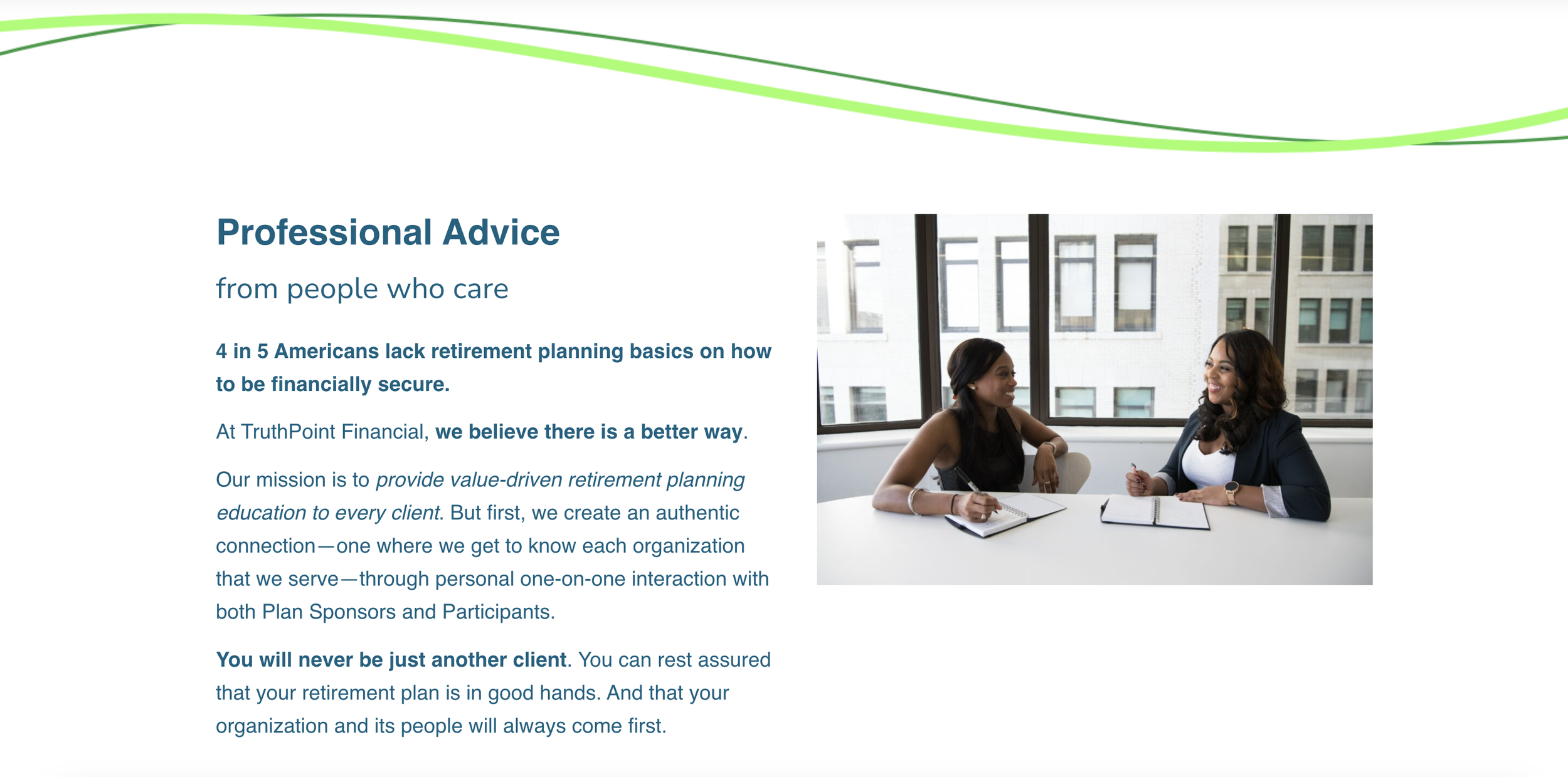 Two women sitting across from each other at a table in a bright office with large windows, having a discussion, with notepads and pens in front of them.