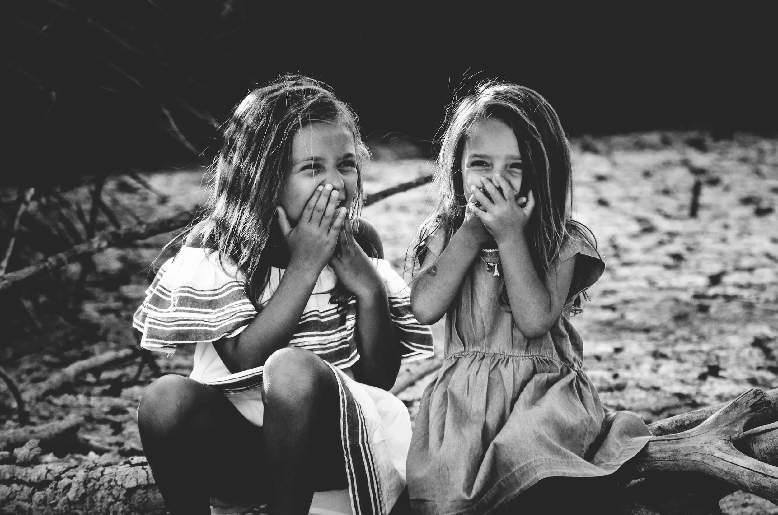 Two young girls sitting outdoors, covering their mouths, smiling and giggling, in black and white.  Learning how to reduce and treat stuttering symptoms