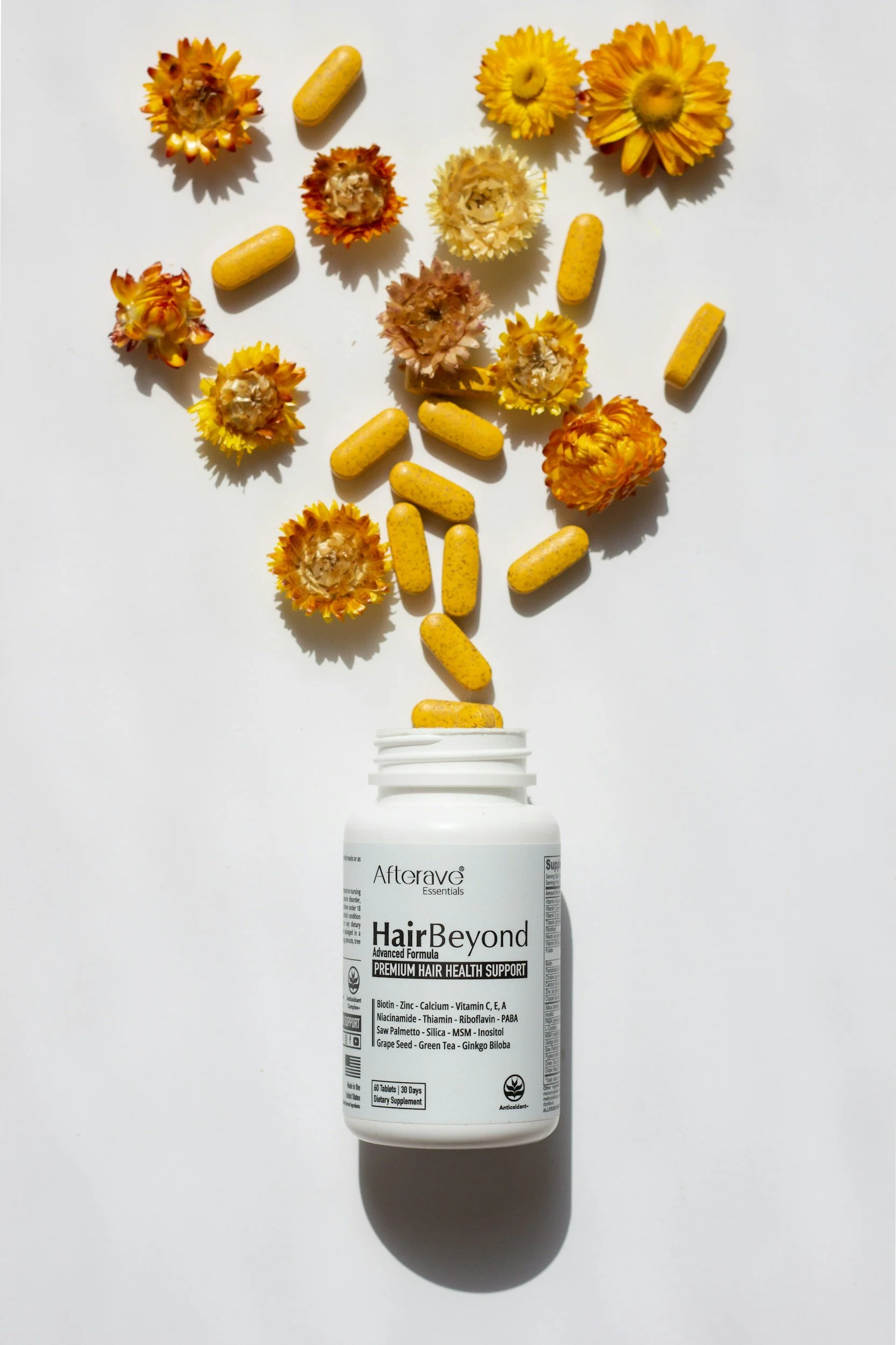 A white bottle of HairBeyond supplement pills with yellow flowers and yellow capsules arranged above it, creating a floral pattern.