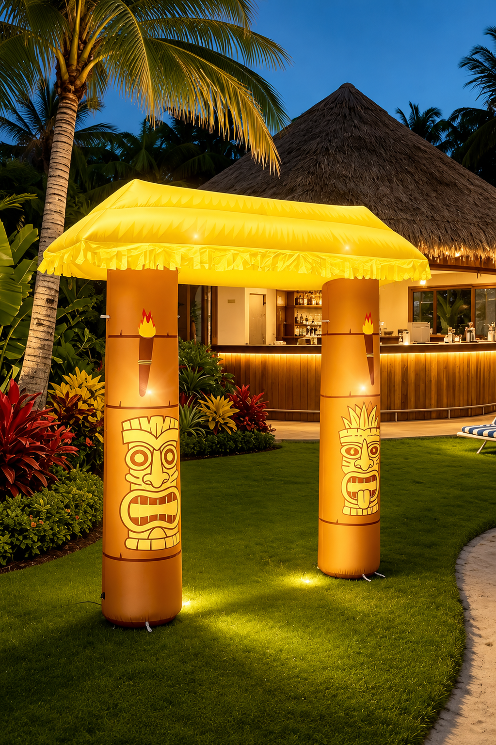 Illuminated inflatable tiki hut arch with carved tiki columns and a glowing thatched roof displayed in front of a tropical resort at sunset, perfect for luaus, Hawaiian parties, beach events, and tropical-themed celebrations.