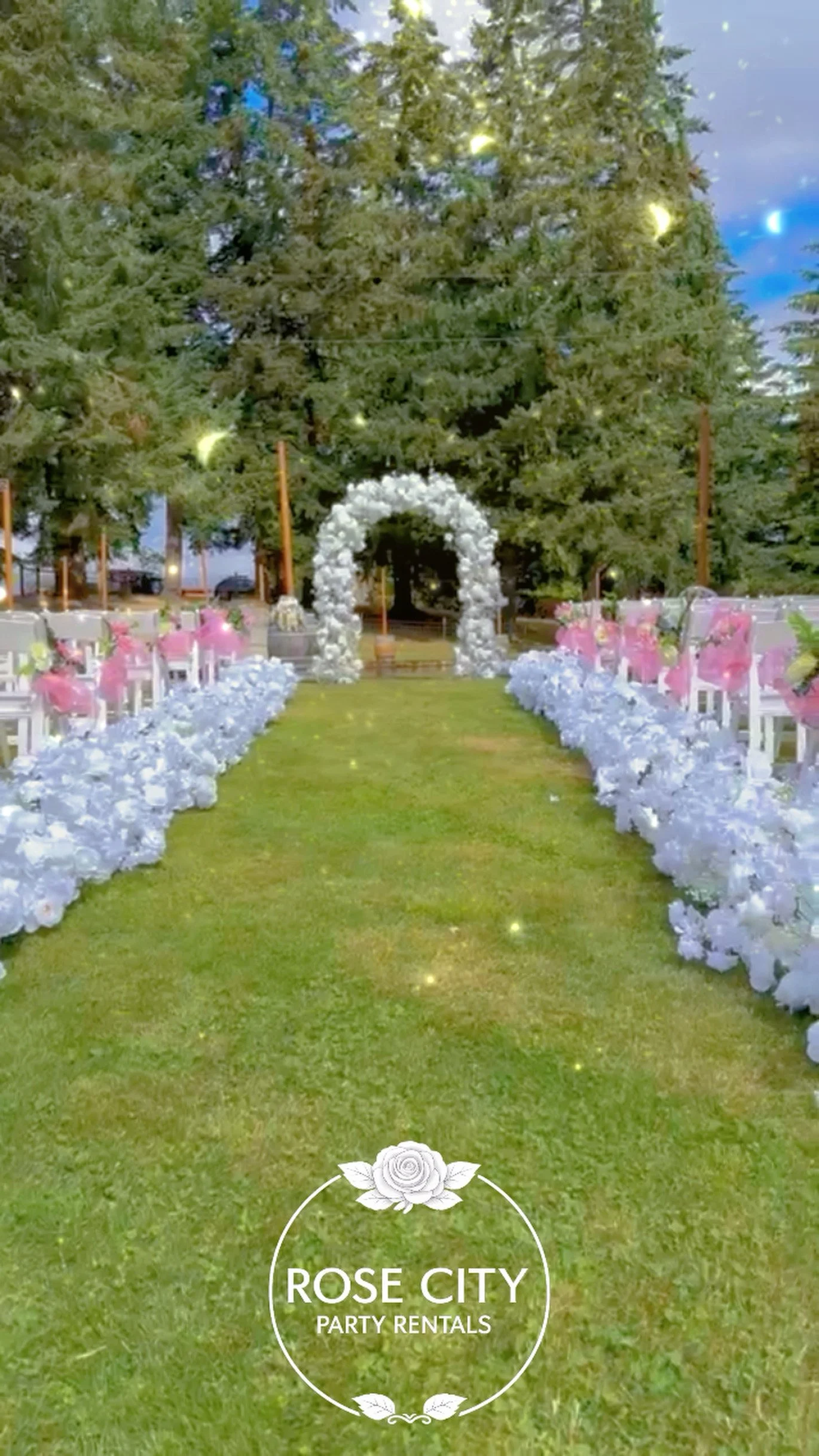 Outdoor wedding ceremony featuring a white floral arch, full white floral aisle arrangements, white guest chairs with pink floral decorations, and towering evergreen trees creating a romantic woodland setting.