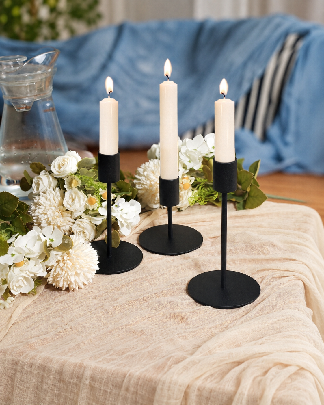 Three matte black candlestick holders of varying heights with lit ivory taper candles on a cream tablecloth beside white flowers.