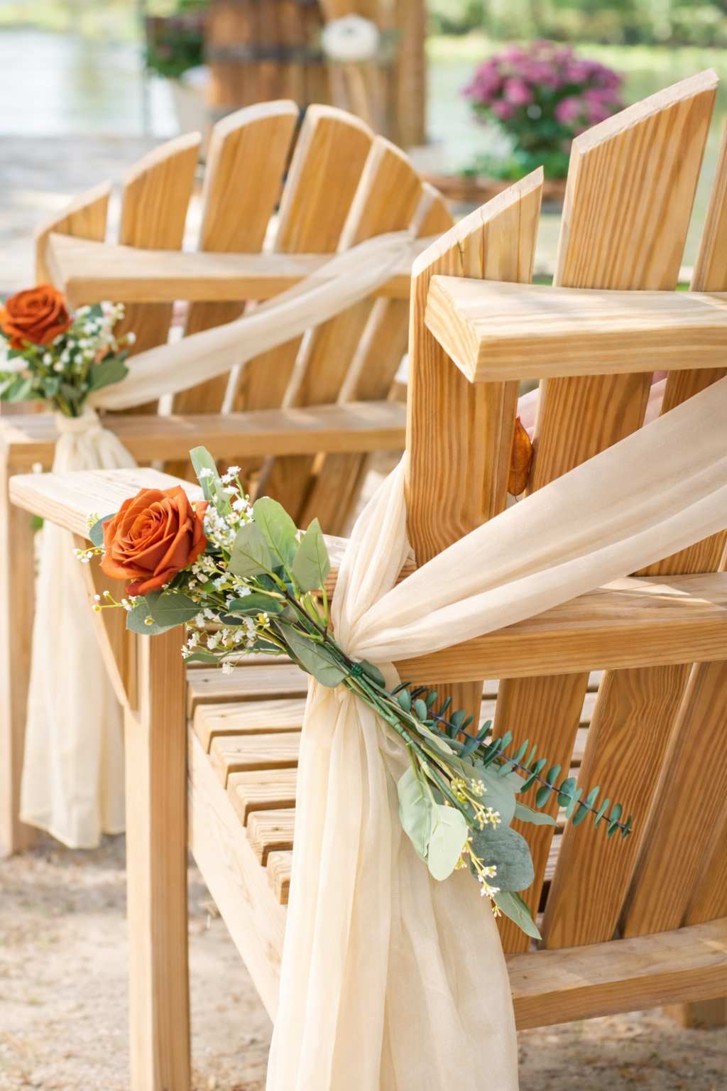 Wooden ceremony chairs decorated with soft cream chiffon sashes and terra-cotta orange roses, accented with greenery and delicate white filler flowers, styled for an elegant outdoor wedding aisle.
