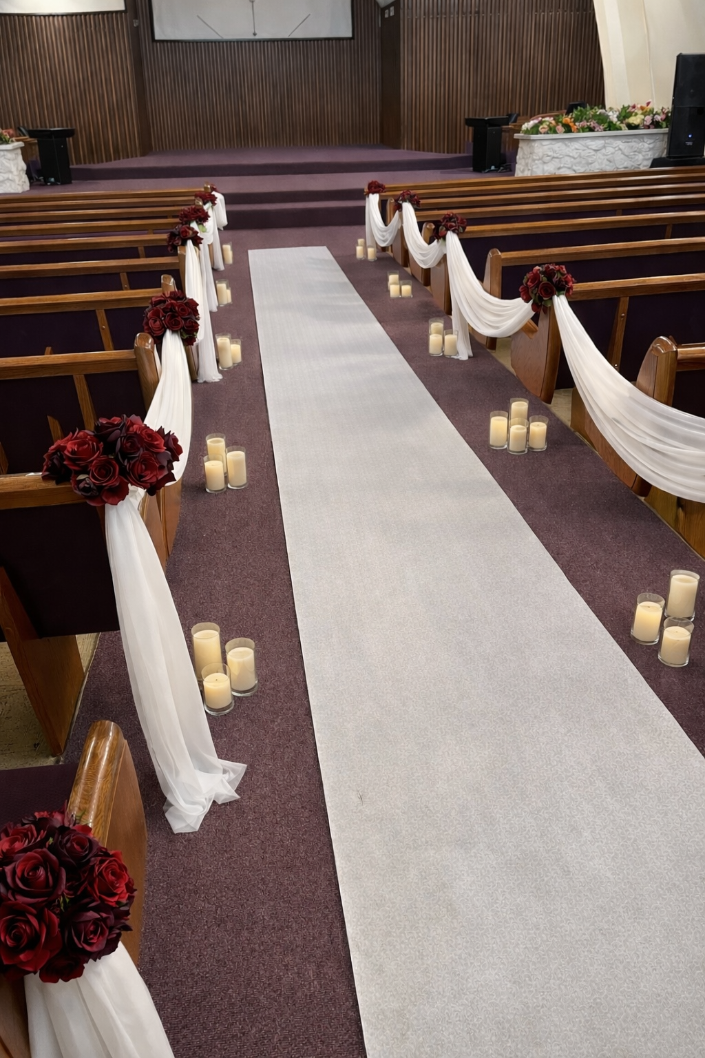 Wedding aisle with white runner, ivory draping, burgundy rose arrangements, and pillar candles lining church pews for a formal ceremony setup.