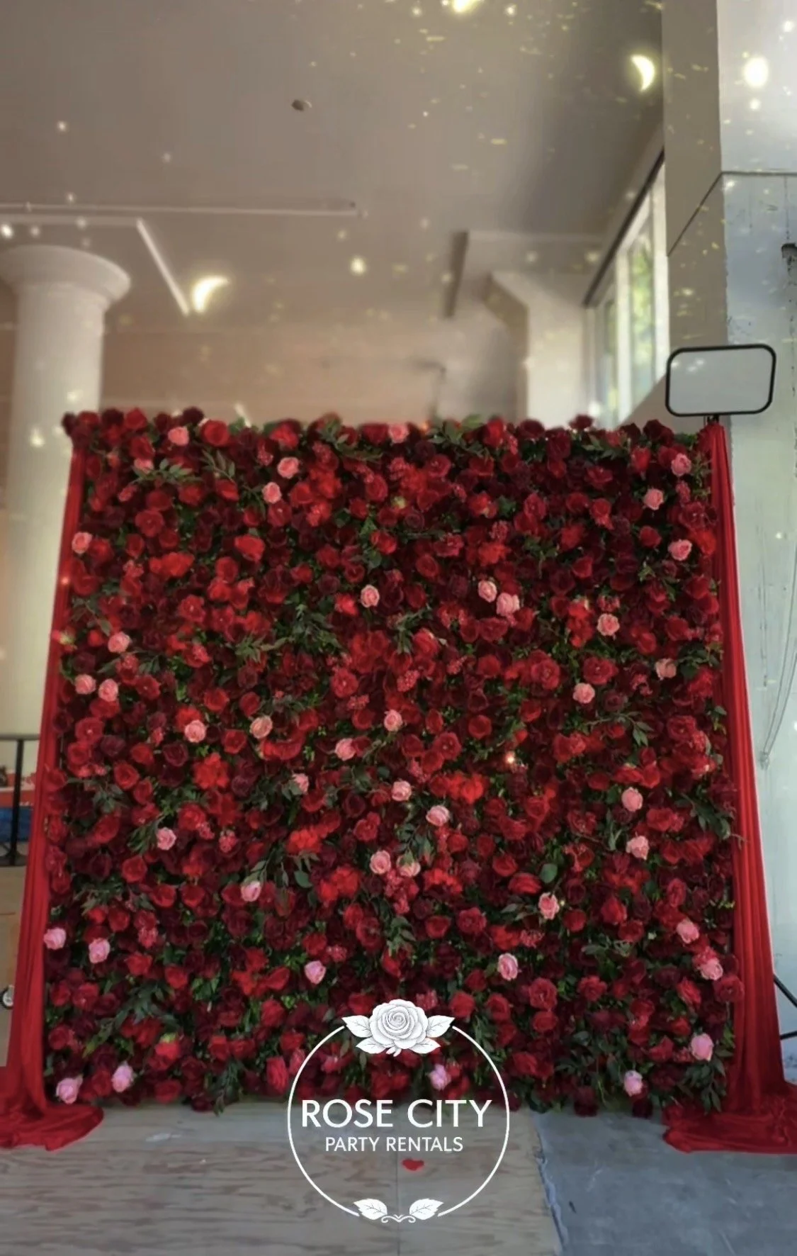 8’ x 8’ artificial floral backdrop wall featuring burgundy, red, and blush roses with red drapes and an LED quote sign, designed as an elegant event photo backdrop.