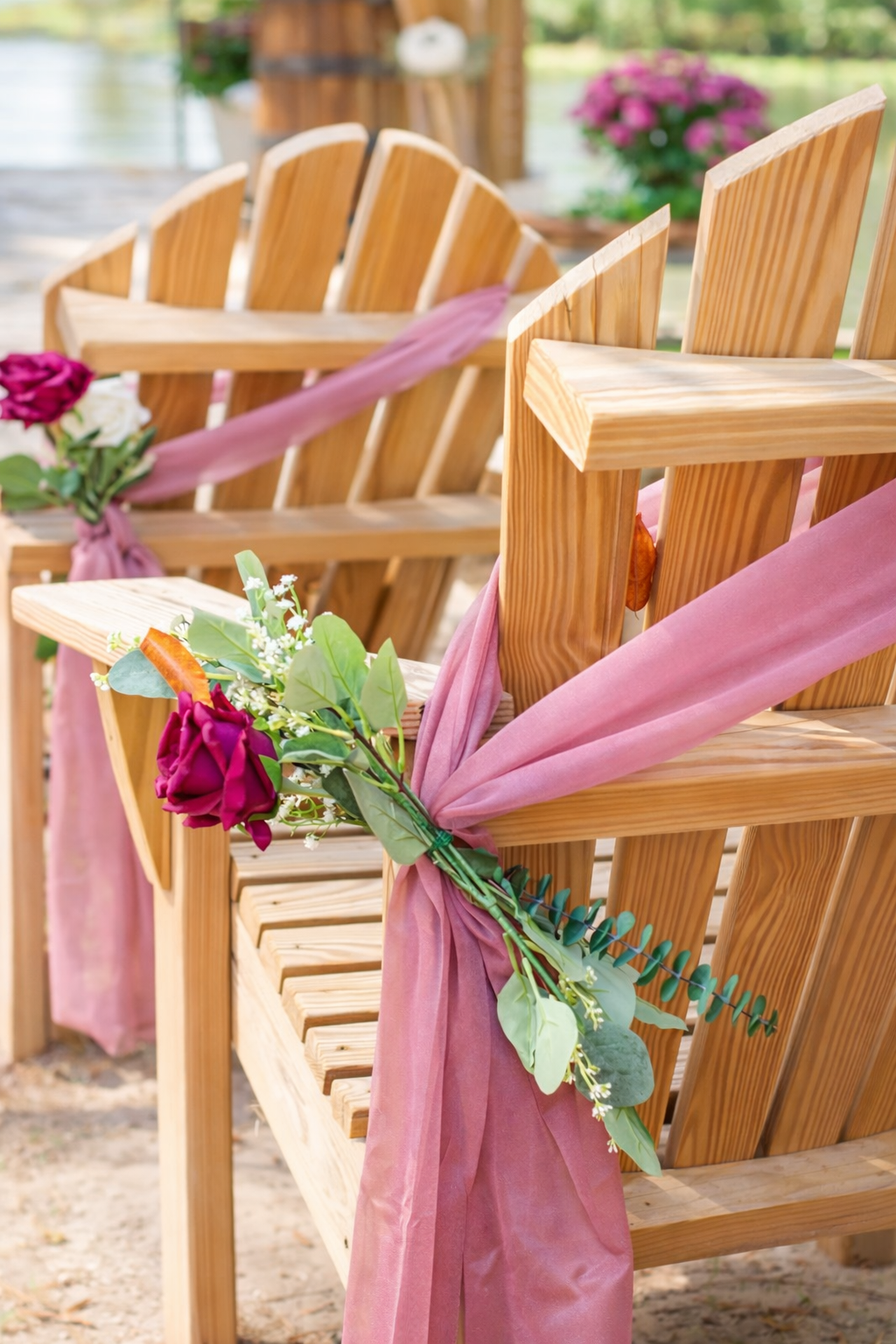 Wooden ceremony chairs decorated with dusty rose chiffon sashes and burgundy roses, accented with greenery and delicate white filler flowers, styled for a romantic outdoor wedding aisle setting.