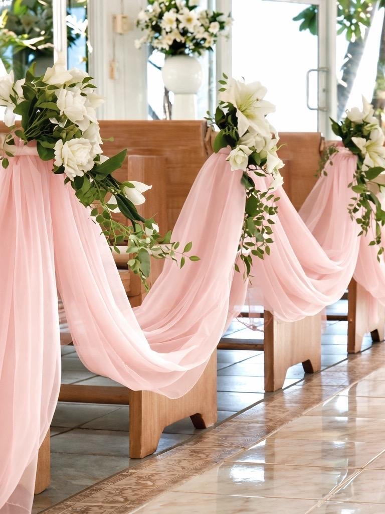 Blush pink chiffon aisle draping with white floral arrangements and greenery on wooden church pews for a romantic wedding ceremony.