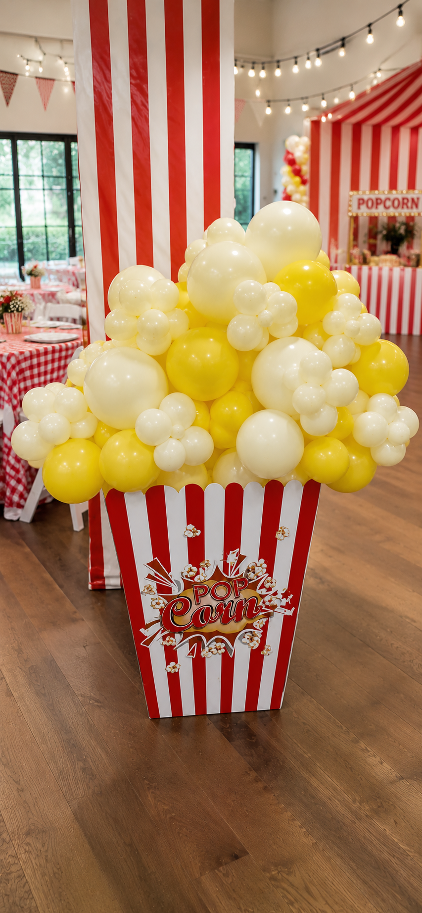 Red-and-white popcorn box balloon display overflowing with clustered white and yellow balloons, styled to resemble popcorn, showcased at a festive carnival-themed party with striped décor and a popcorn concession stand in the background.