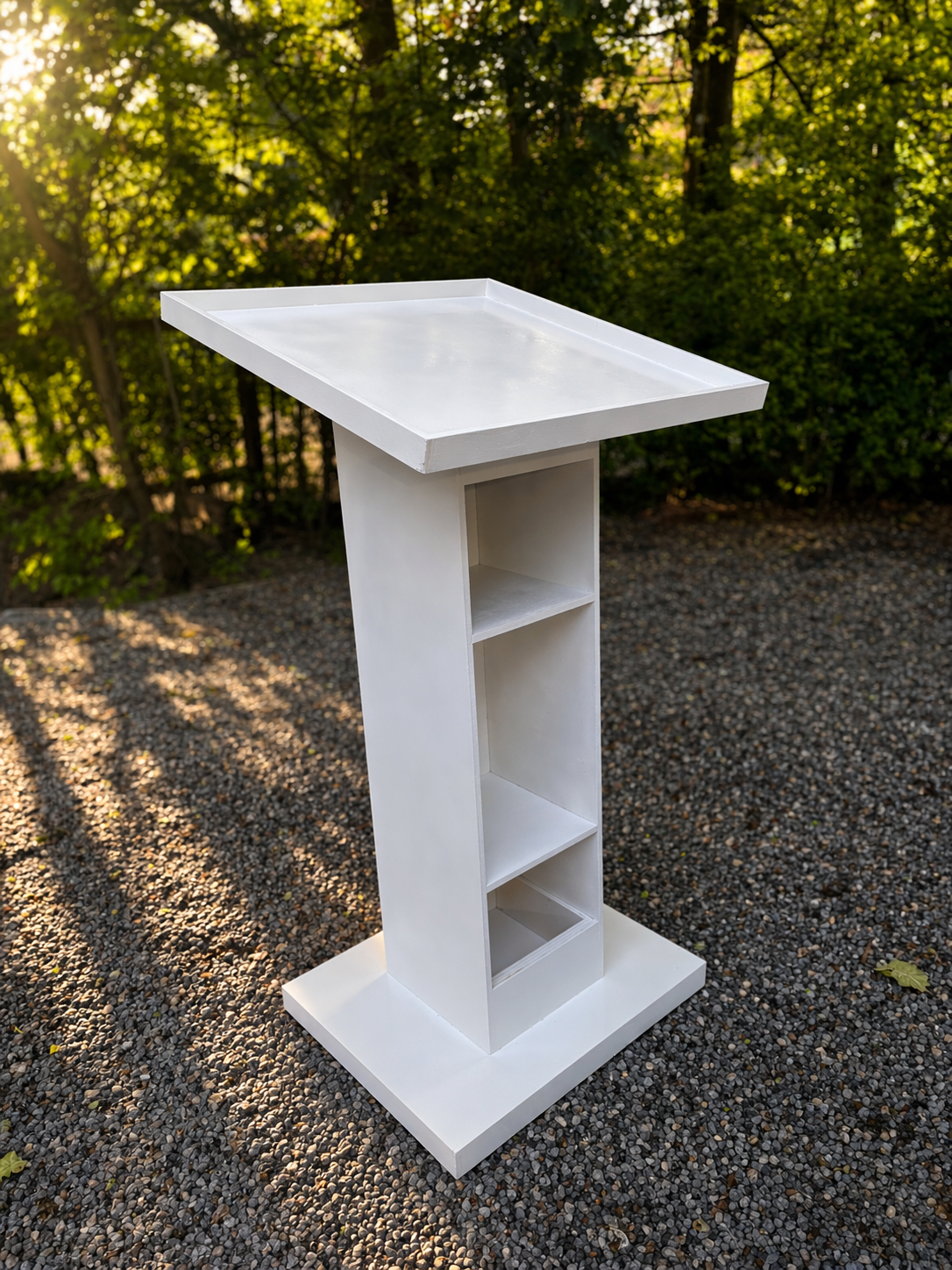 White modern podium with a slanted top and built-in shelves, displayed outdoors on a gravel surface with trees in the background.
