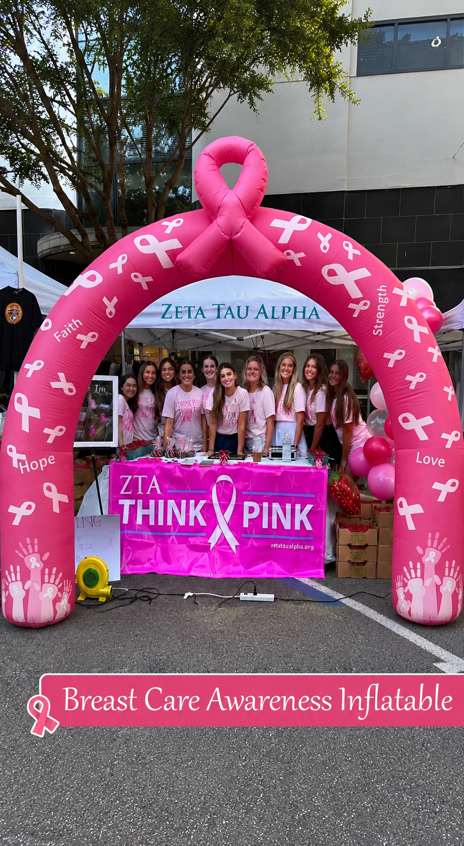 Large pink breast cancer awareness inflatable arch with a ribbon topper framing a fundraising booth staffed by volunteers in matching pink shirts, decorated with balloons and a “Think Pink” banner at an outdoor community event.