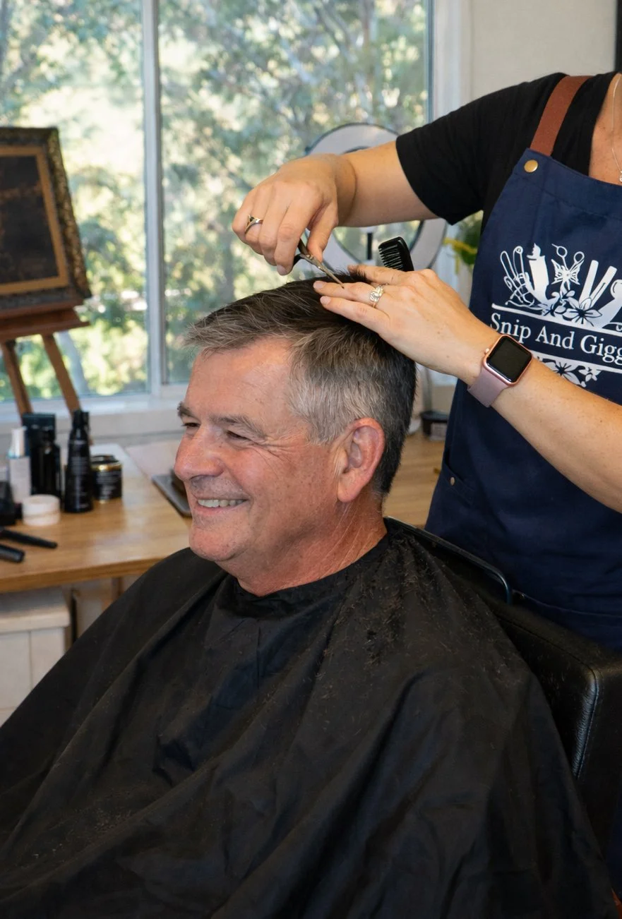 A man getting a haircut at a salon, smiling while a hairstylist cuts his hair with scissors. The man is sitting in a chair, wearing a black cape, and the stylist is wearing a navy apron and smartwatch. The background shows a window and salon supplies.