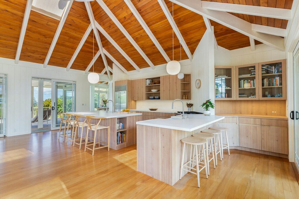 Open kitchen with wooden cabinets and white countertops, kitchen island with bar stools, large windows, and a high wooden ceiling with exposed beams.