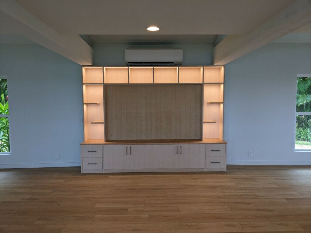 Empty room with built-in wooden shelving and cabinets, two windows showing greenery outside, and a mounted air conditioning unit.