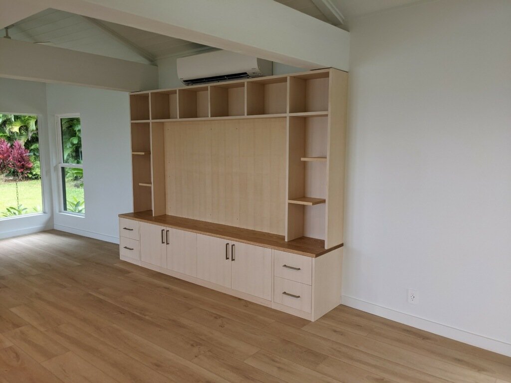 Empty room with wooden flooring, a large built-in wooden shelving unit, and two windows looking outside to green plants.