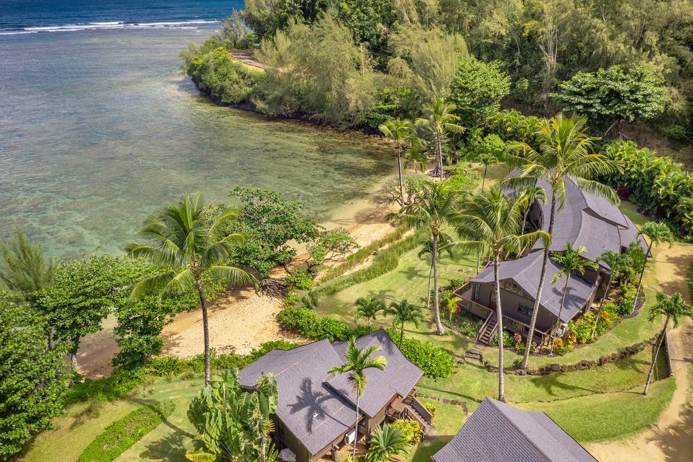 Aerial view of a tropical beachside property with palm trees, green lawns, and multiple houses near the shoreline with clear water and lush greenery.