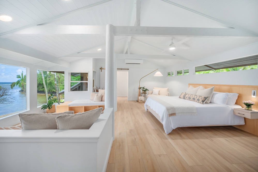 Bright, airy bedroom with white walls, light wood flooring, a large bed with white bedding, and windows with views of palm trees and water.
