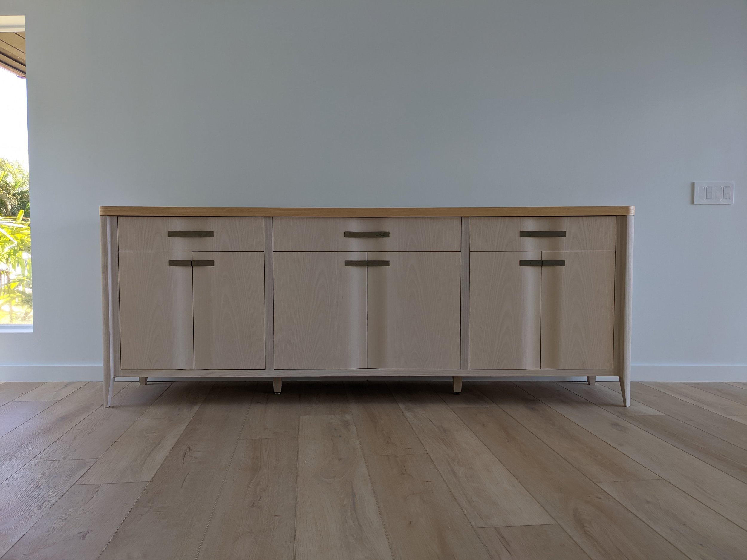 Minimalist wooden sideboard with four doors, placed against a white wall in a bright room with wooden flooring, with a window showing greenery outside.
