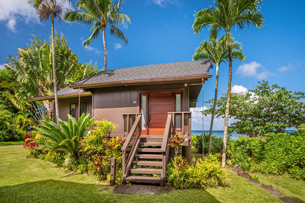 A small wooden house on stilts surrounded by tropical plants and palm trees, with a view of the ocean in the background under a blue sky.