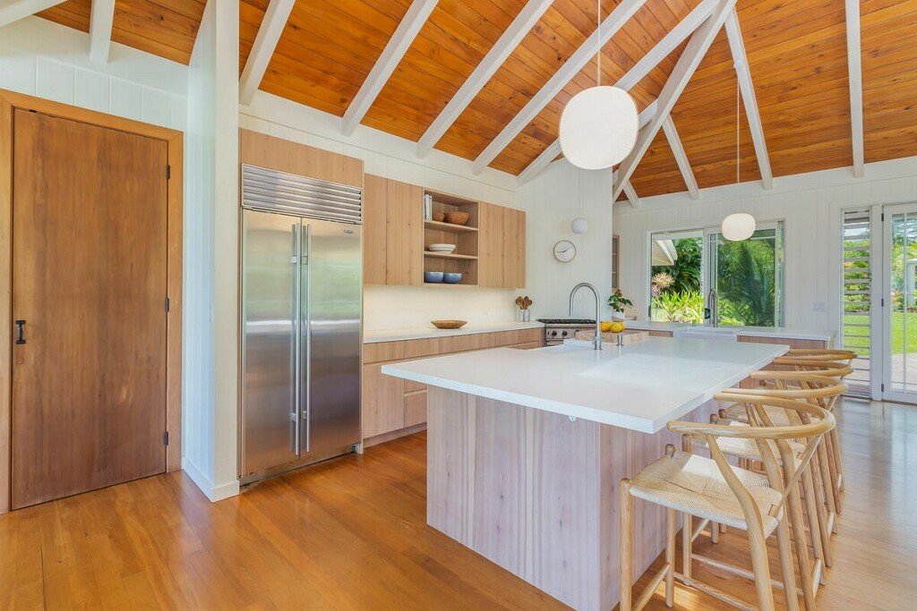 Modern kitchen with wooden vaulted ceiling, white walls, stainless steel refrigerator, open shelving, a large kitchen island with beige bar stools, and large windows showing greenery outside.