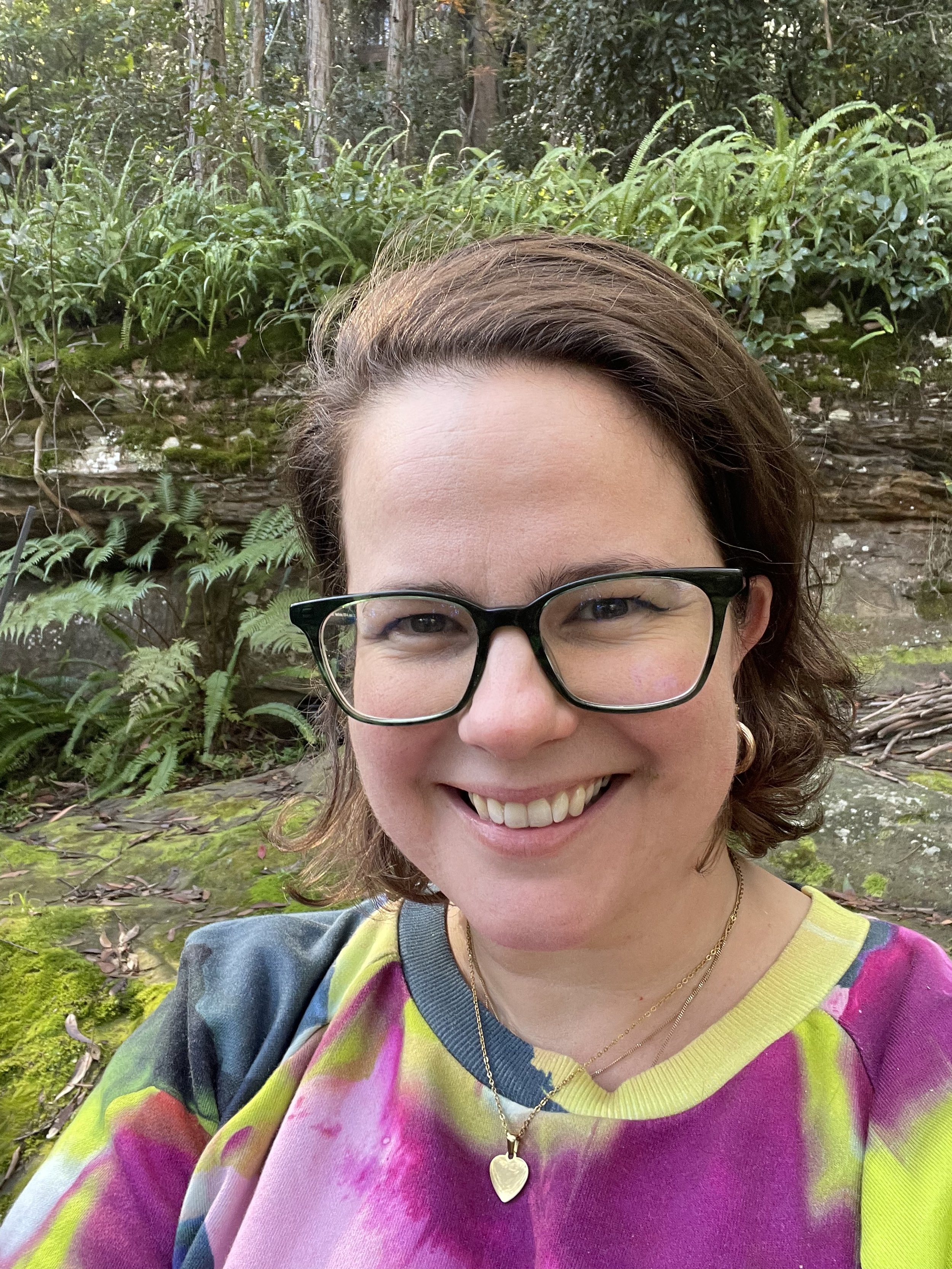A woman with short brown hair, wearing black glasses and a colorful tie-dye shirt, is smiling outdoors in front of a green, wooded background. She has gold necklaces, one with a heart-shaped pendant.