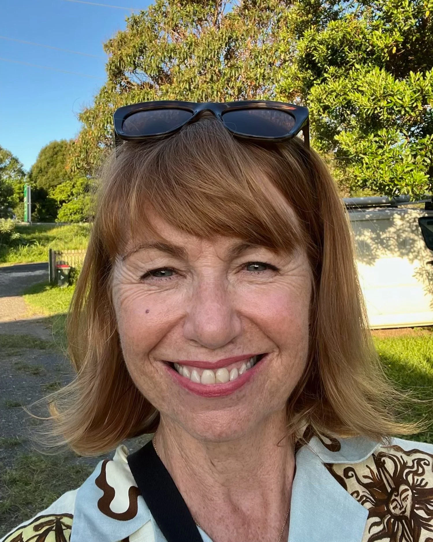 A smiling middle-aged woman with red hair and sunglasses on her head outdoors on a sunny day, with trees and a blue sky in the background.