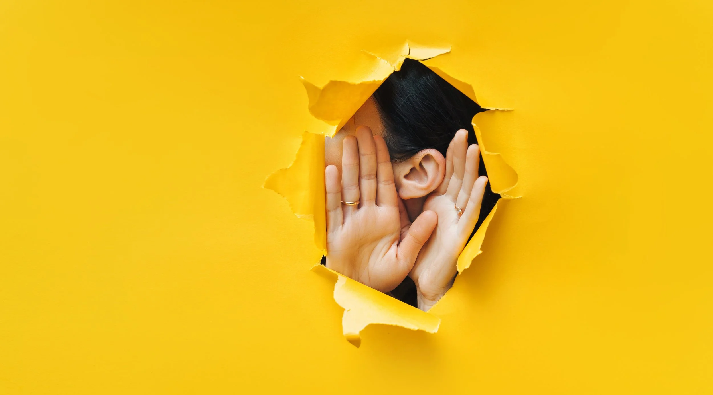 Person peeking through a torn yellow paper wall, covering their face with both hands.