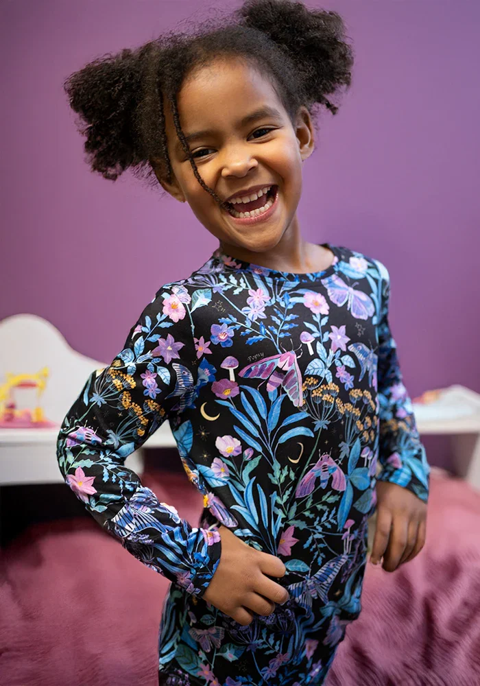 Smiling young girl with curly hair wearing a colorful floral patterned outfit, standing in a bedroom with a purple wall in the background.