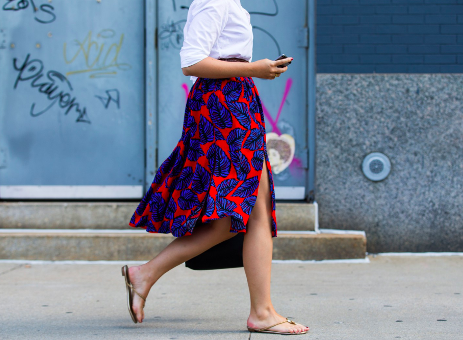 A person walking on the sidewalk wearing a white top, a colorful skirt with a blue and red leaf pattern, and flip-flops, holding a smartphone in their hand.