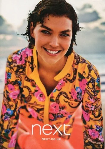 A young woman with short dark hair smiling on a beach, wearing a colorful, floral patterned jacket.