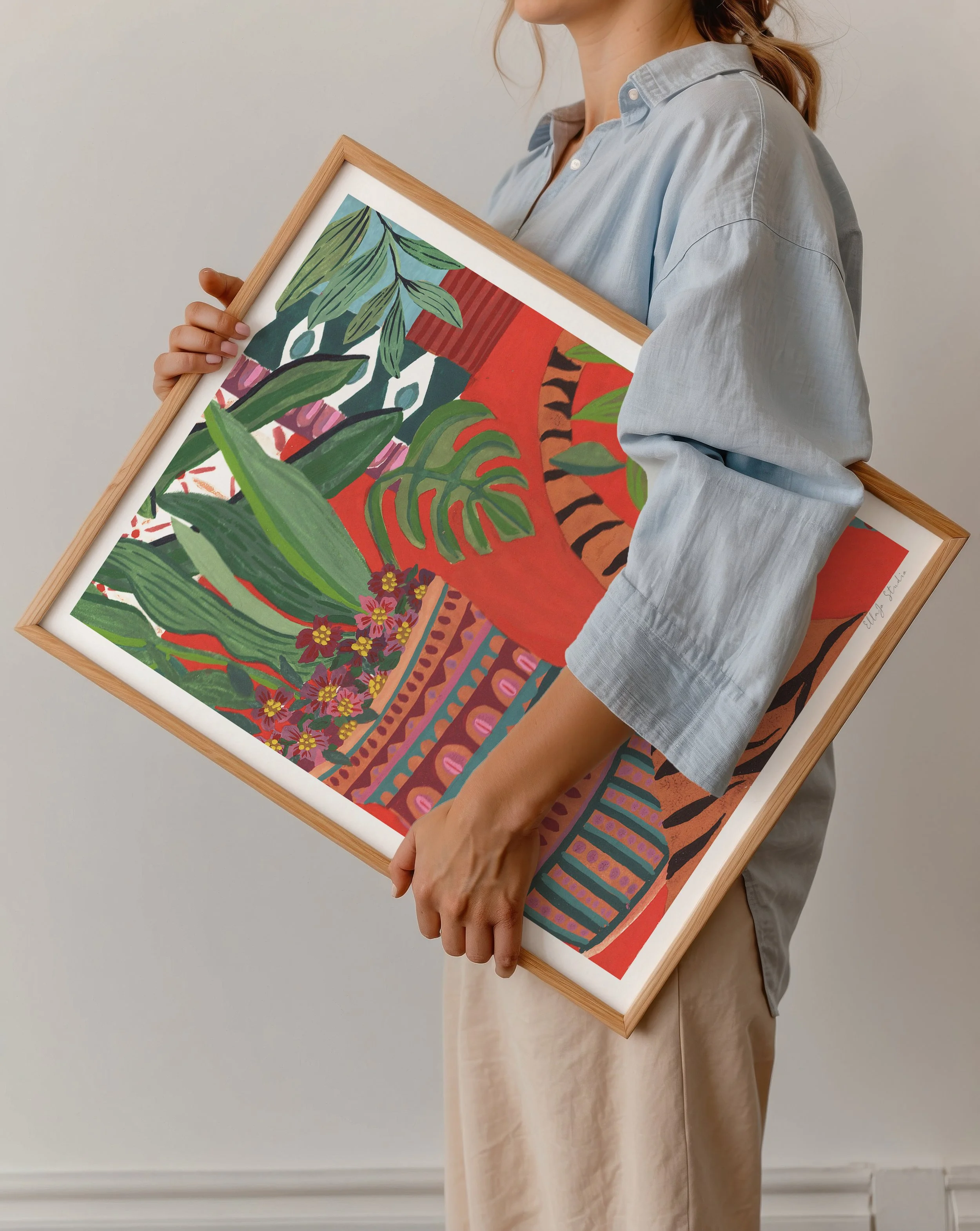 Person holding a framed colorful tropical-themed artwork featuring lush green leaves, vibrant flowers, and bold patterns.