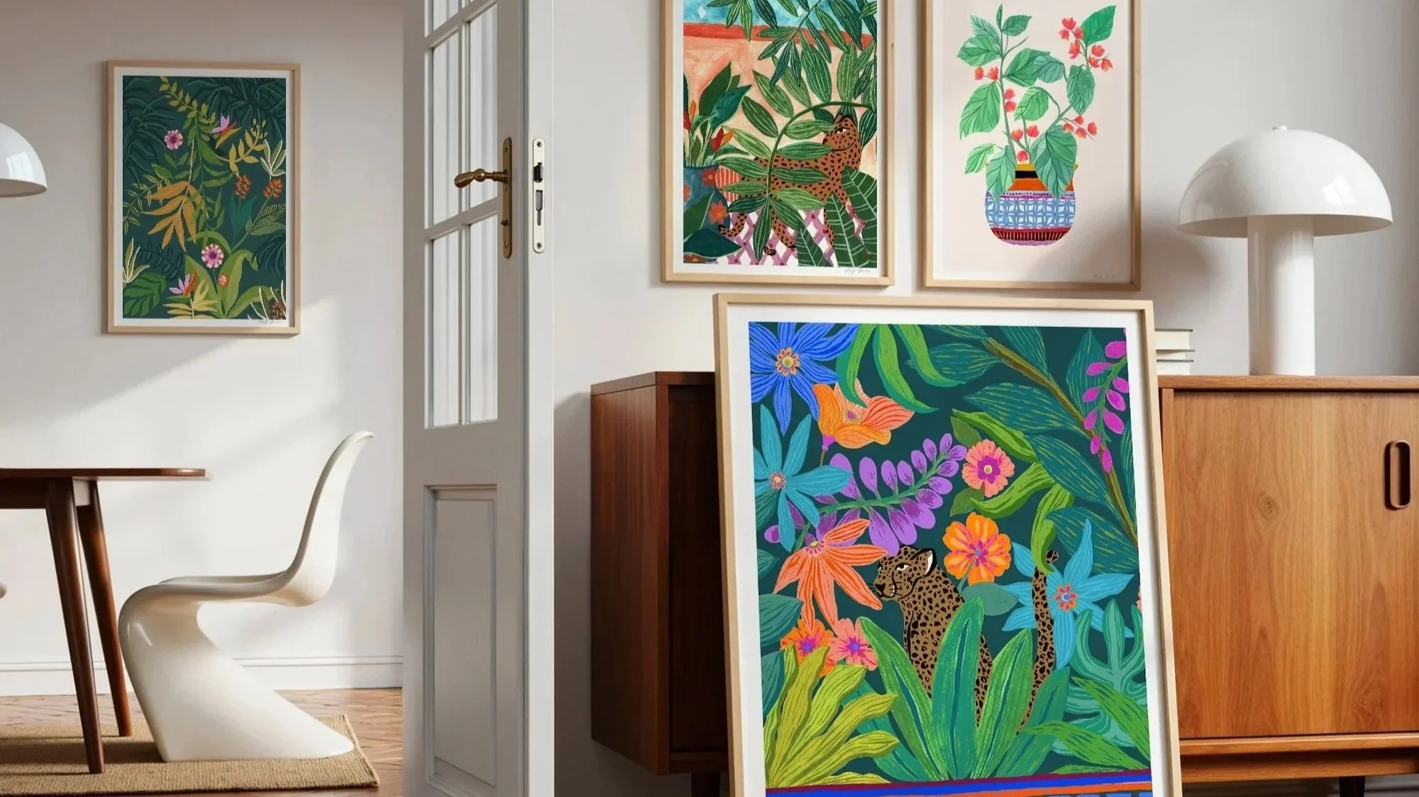 An interior room decorated with vibrant jungle-themed artwork, including a large framed painting of two leopards amongst lush greenery and colorful flowers leaning against a wooden cabinet, and three smaller framed illustrations of plants and animal life on the wall. There is a white table lamp on a wooden sideboard, a white curved chair, and a glass-paneled door.