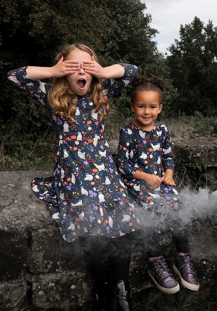 Two young girls in matching dark dresses with white unicorns and colorful flowers, sitting on rocks outdoors. One girl has her eyes covered and mouth open, while the other is smiling.