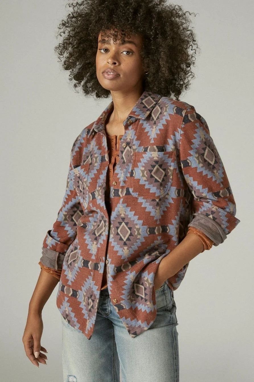 A woman with curly hair wearing a patterned button-up shirt over a brown top and light blue jeans, standing against a plain background.
