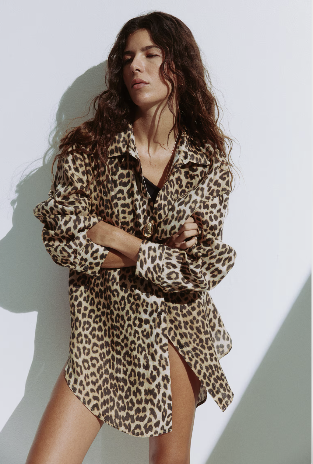 H&M lookbook editorial, a woman with wavy brown hair wearing a leopard print shirt by EllaJo Studio with her arms crossed, standing against a white wall with a shadow.