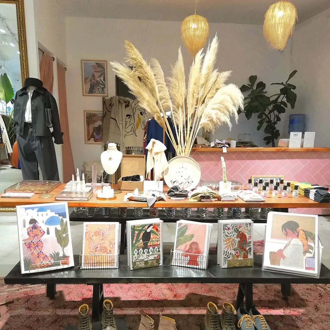Display table with artwork prints in wire baskets, shoes at the front, and various decor items including pampas grass in a vase, jewelry, and fabric samples at a boutique.