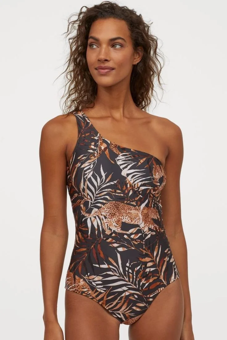A woman with curly hair wearing a one-shoulder swimsuit with a jungle print featuring leopard and tiger images and tropical leaves.