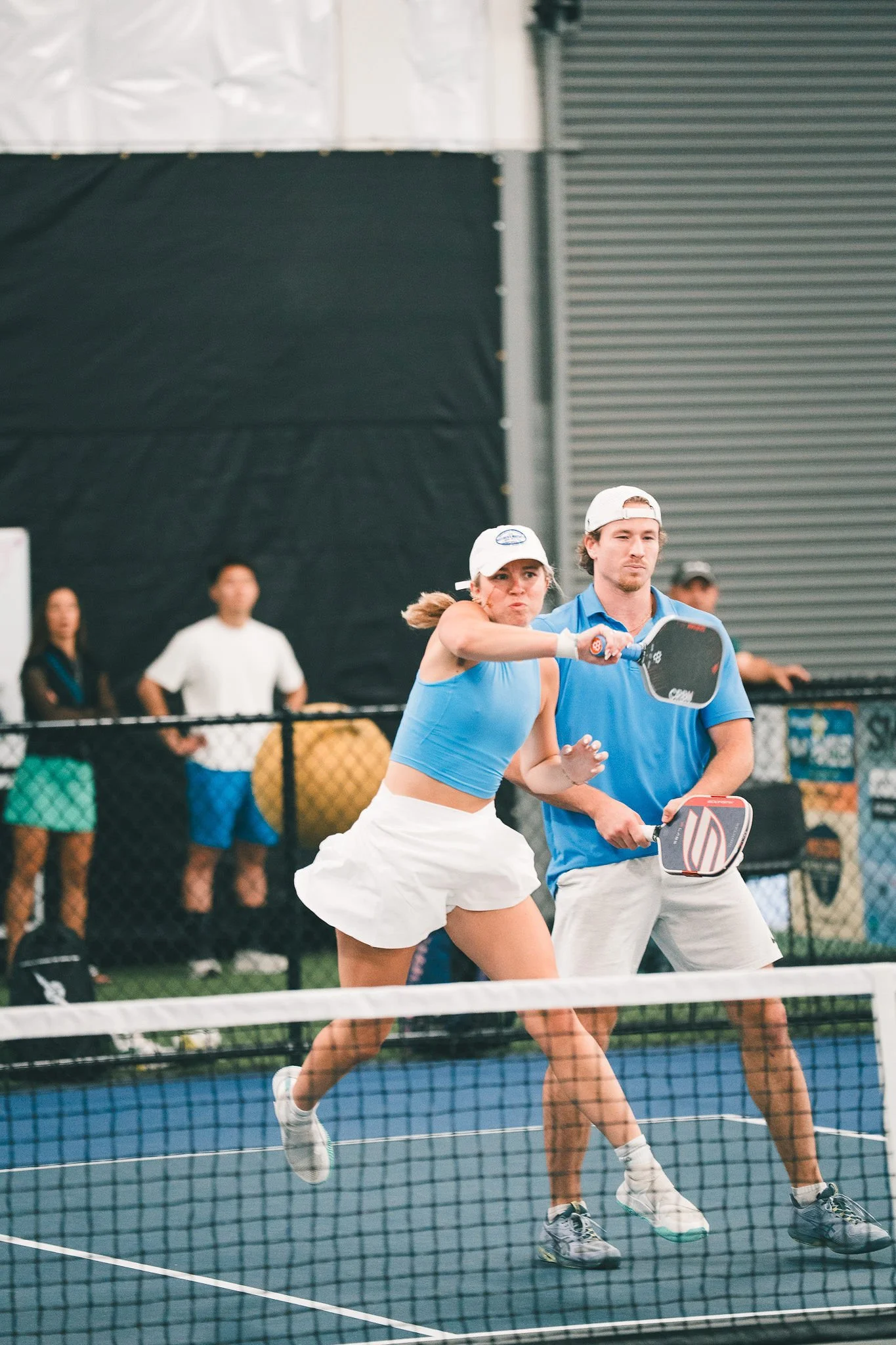 Two people playing pickleball on an indoor court, with a woman in a blue tank top and white skirt hitting the ball and a man in a blue shirt and white shorts holding a paddle. There are spectators behind a fence in the background.