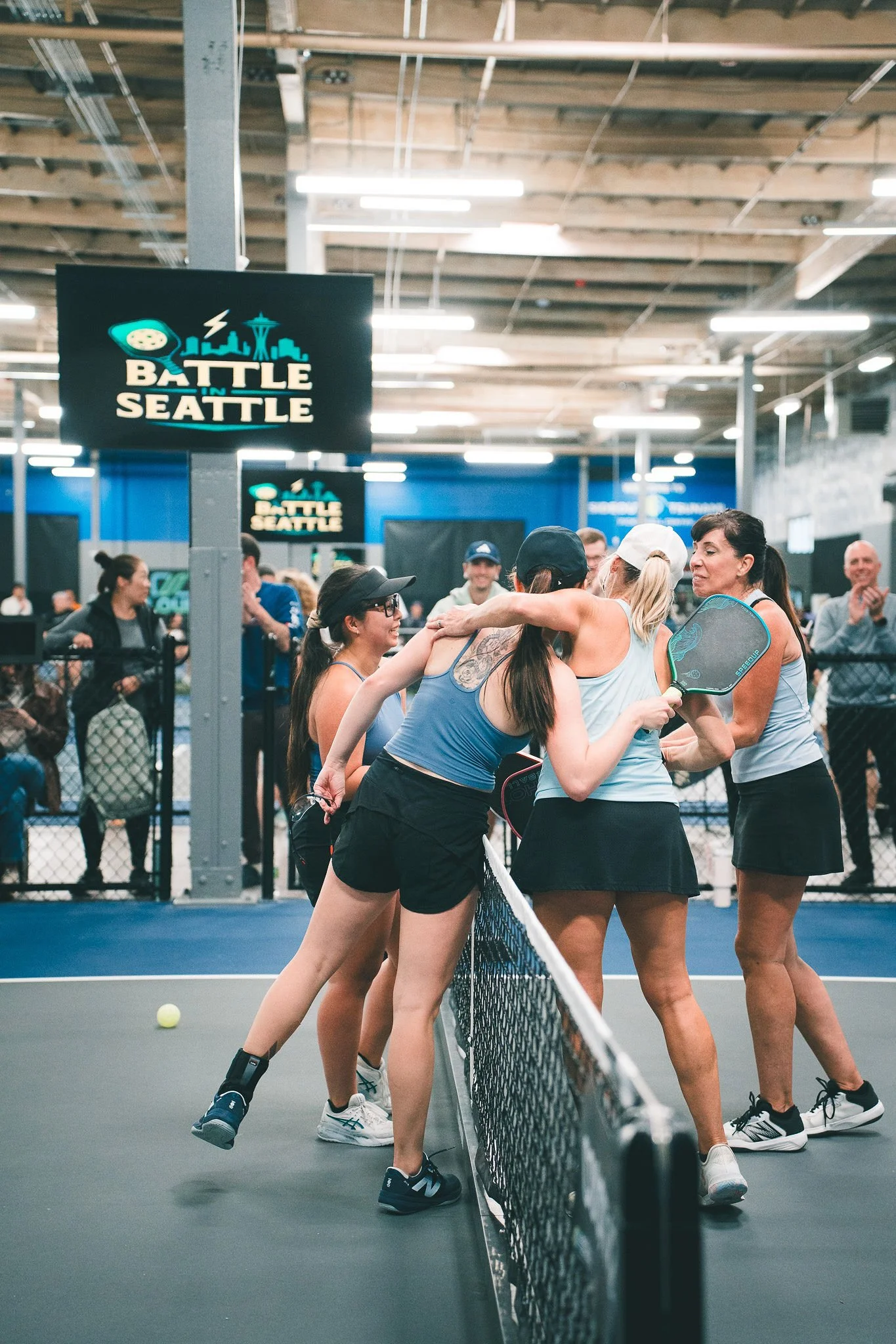 Women on a tennis court, some wearing athletic gear including shorts, tank tops, and sneakers, gathered near the net, with some hugging and others holding tennis racquets. The court has a blue surface, and there are signs that read 'Battle in Seattle' on screens hanging from the ceiling. Other people and spectators are visible in the background.