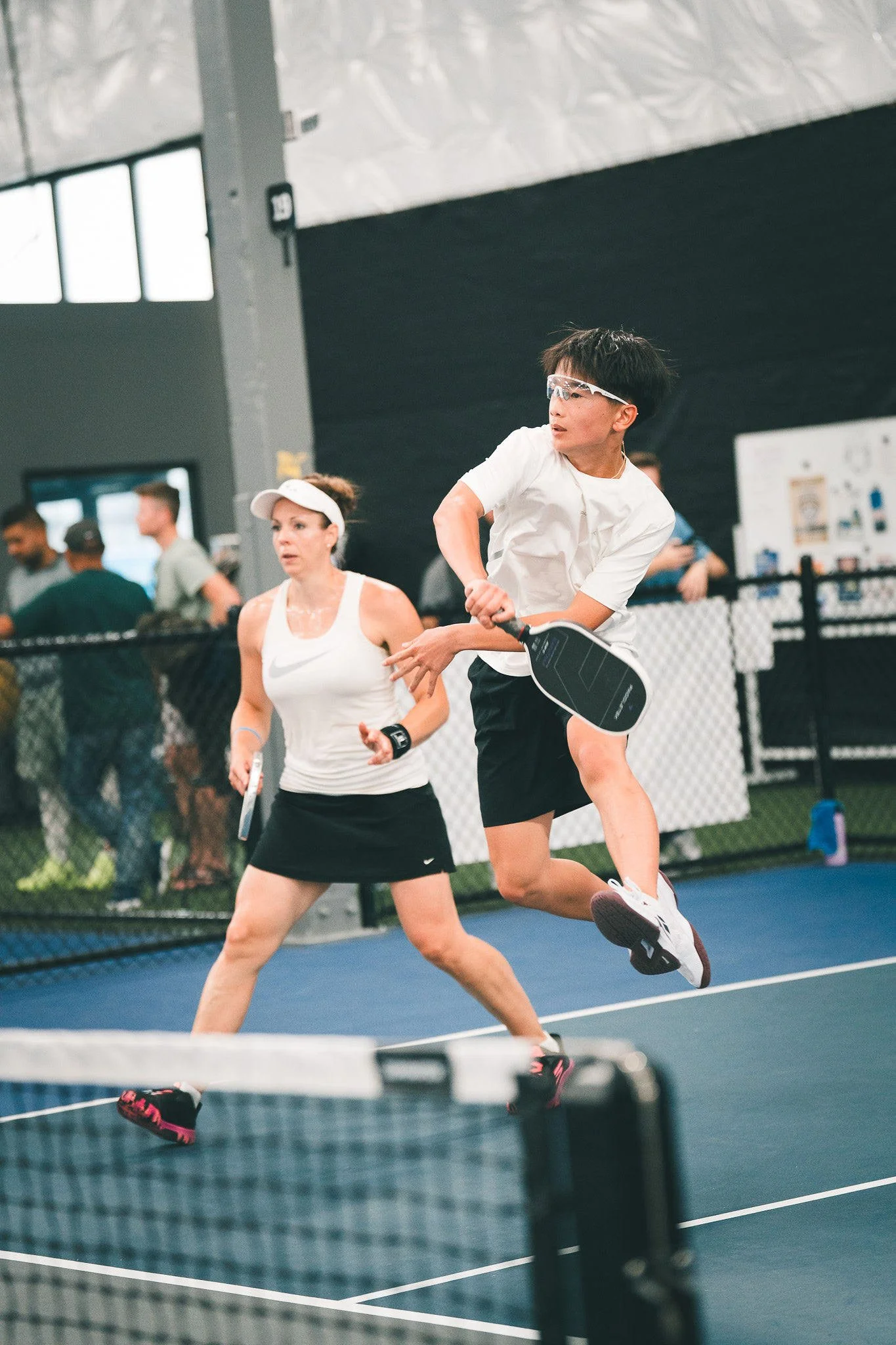 Two people playing pickleball on an indoor court. The person in the foreground is a woman wearing a white cap, a sleeveless white top, and a black skirt, holding a paddle. The person in the background is a young man jumping in the air, wearing glasses, a white shirt, black shorts, and white sneakers, also holding a paddle. There are other people and a black net in the background.